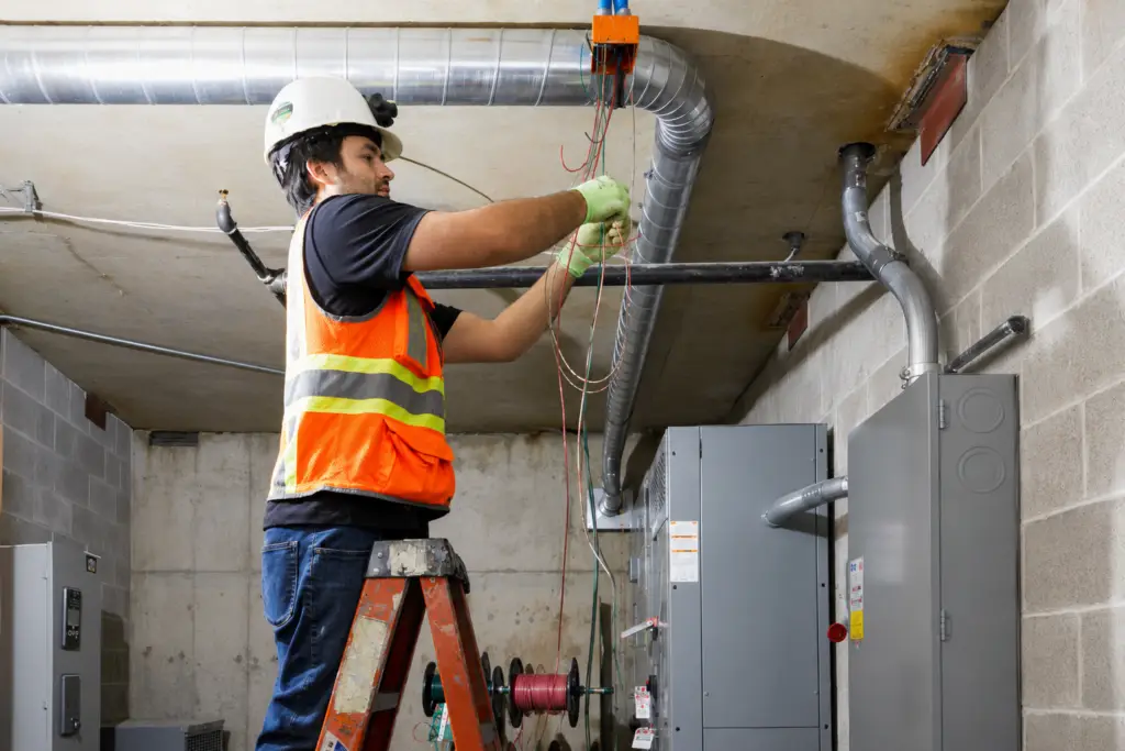 Electrician wearing safety gear installing electrical wiring from a ladder in a commercial mechanical room